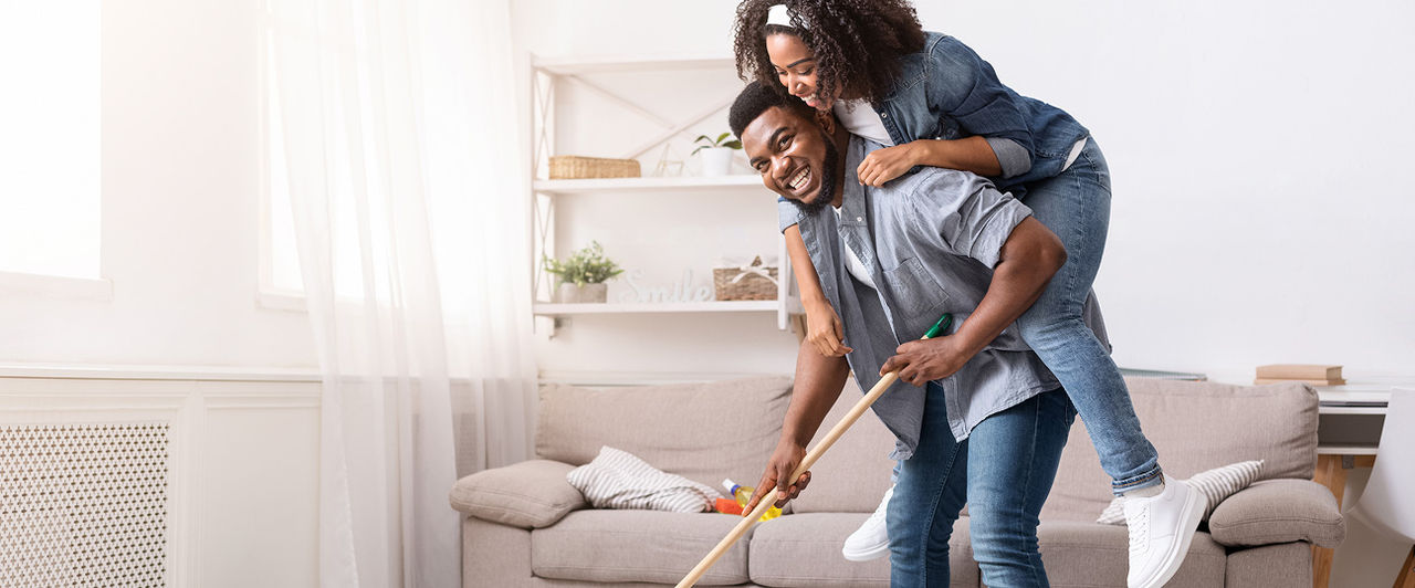 A man gives a piggyback ride to a smiling woman while he holds a mop in a clean room.